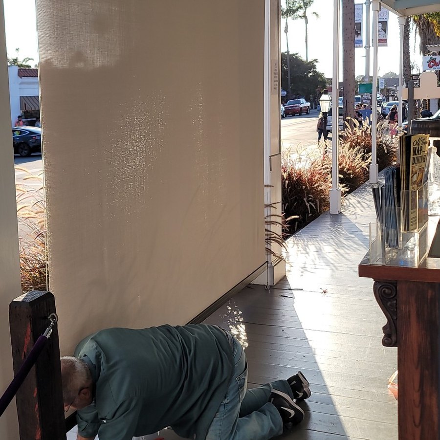 Exterior solar screen lowered across a covered boardwalk, installer fitting the bottom track