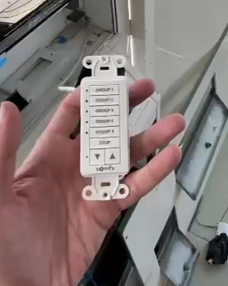 A five-group Somfy wall keypad held in front of an open low-voltage box during installation